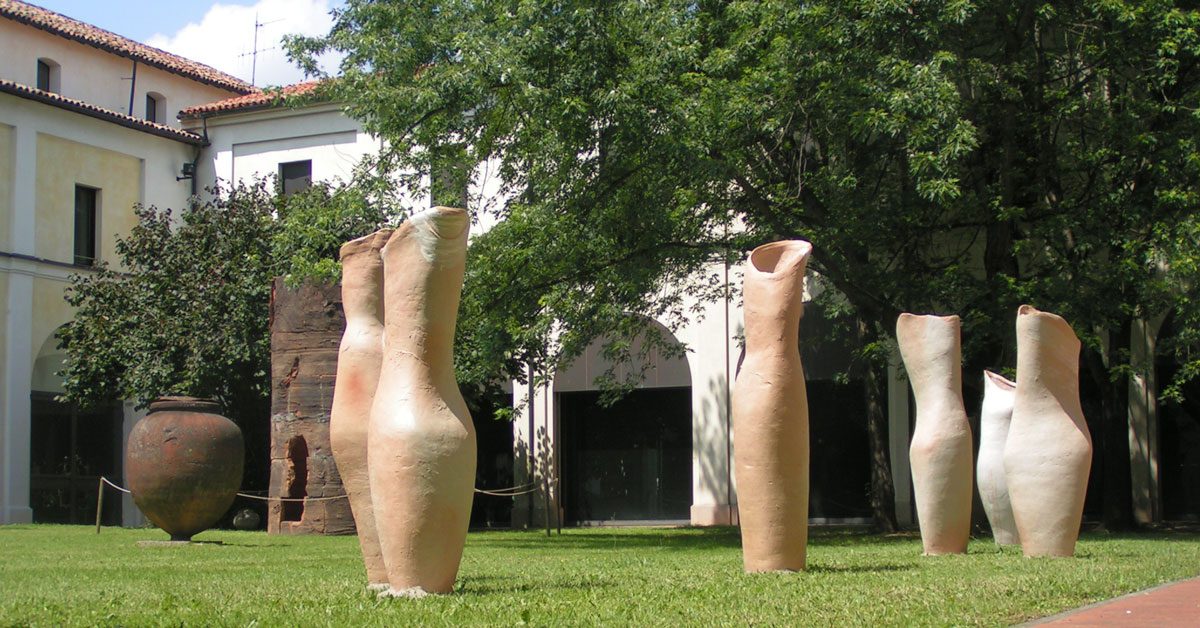 Tall terracotta-coloured vessels on a lawn in a sunlit courtyard
