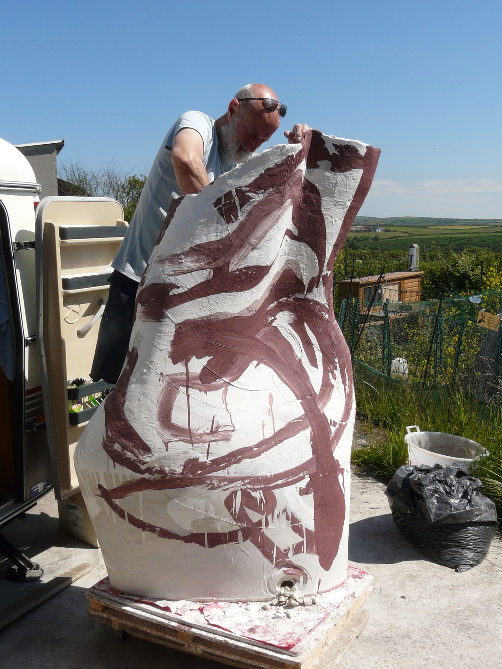 A man leaning over a very large white vessel painted with sweeping maroon marks, outdoors in sunshine